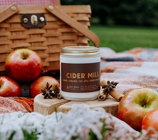 A jar candle labeled Cider Mill next to apples, cinnamon, and a fall blanket