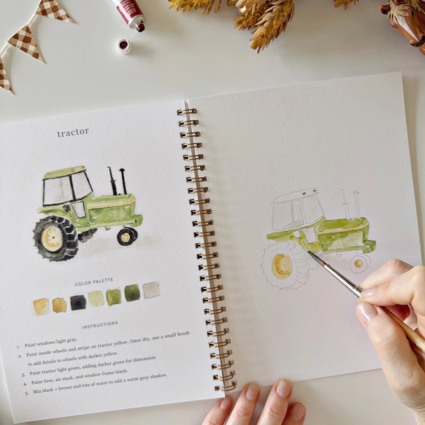 Person drawing a tractor from a watercolor book on a table with paint supplies.