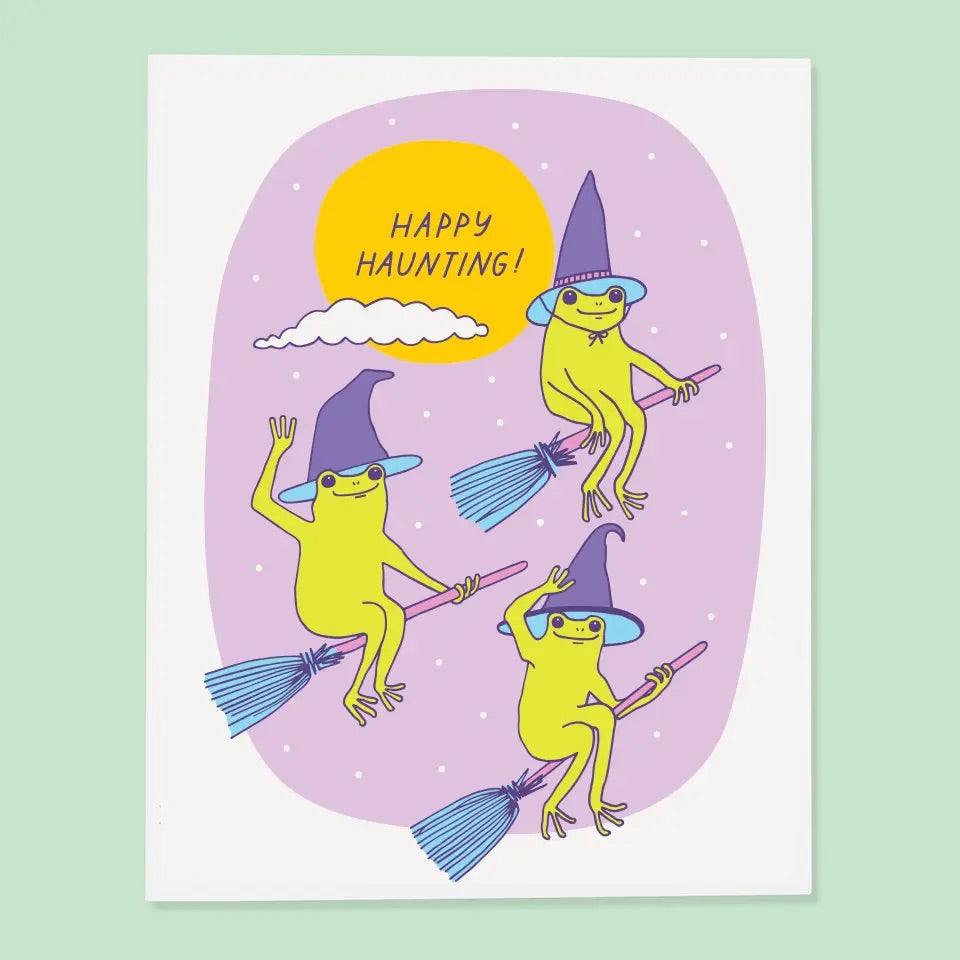 White card with purple background. Illustration of three green frogs on brooms wearing witches hats. Purple text reads “happy haunting!”