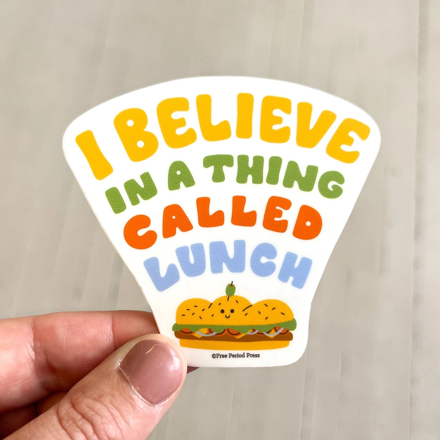 Sticker with white background. Multicolor text "I believe in a thing called lunch" and sandwich illustration