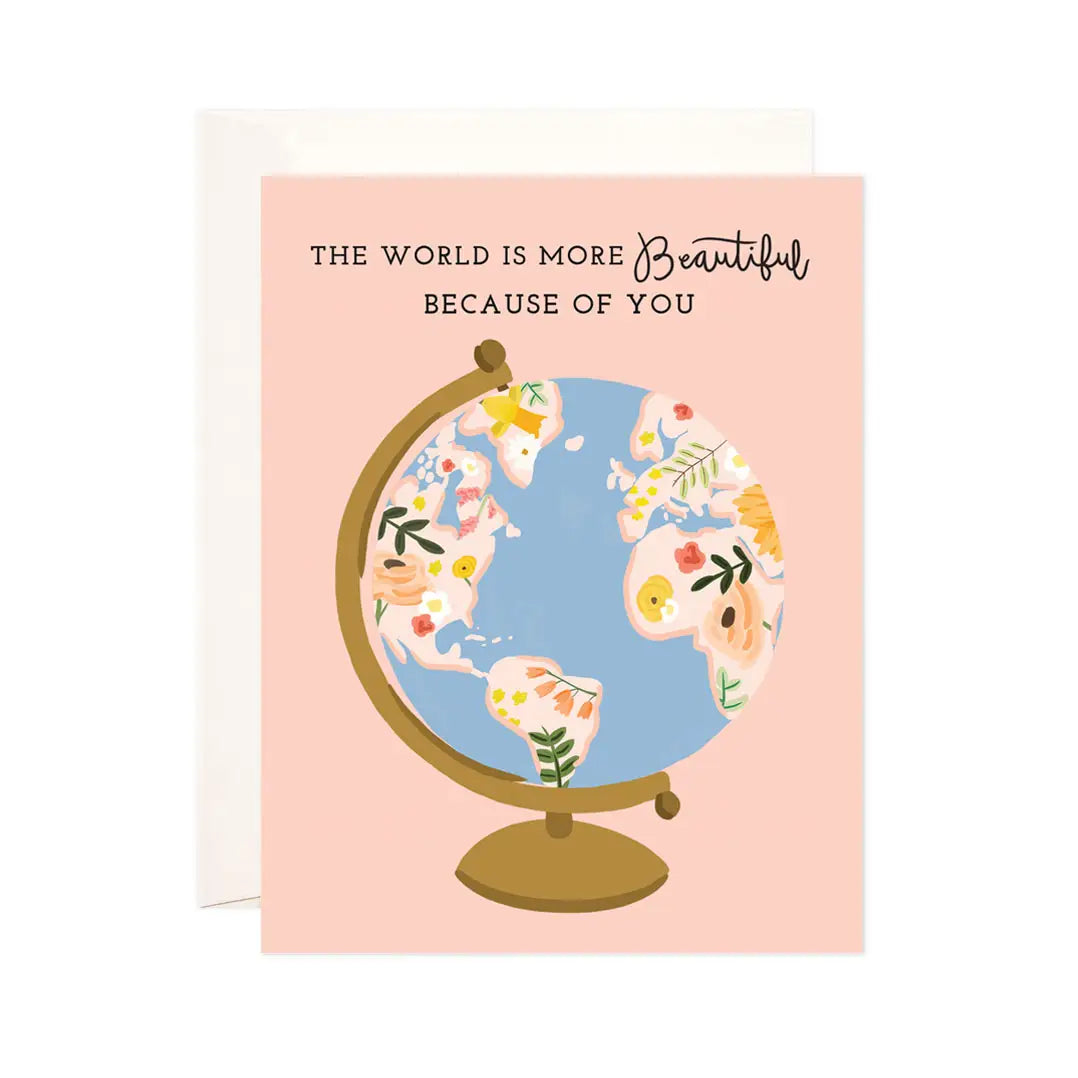 White greeting card with full color pink background. Illustration of globe. Black text “the world is more beautiful because of you”