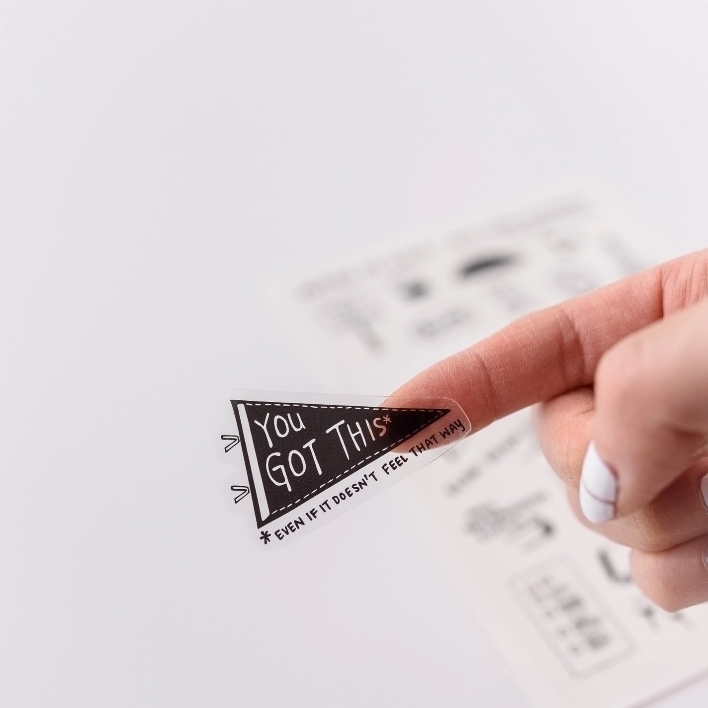 Finger holding a self care sticker in the shape of a pennant. Black background with clear text reads "you got this"