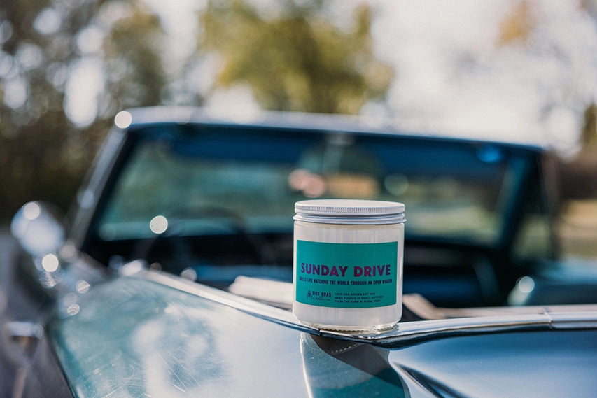 White candle in a clear jar. Blue label. Label text: sunday drive