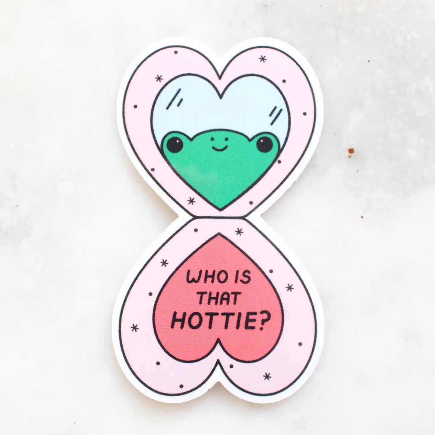 Die-cut sticker in the shape of a heart-shaped open makeup compact. Illustration of a frog looking into a mirror. "who is that hottie" in black text. 