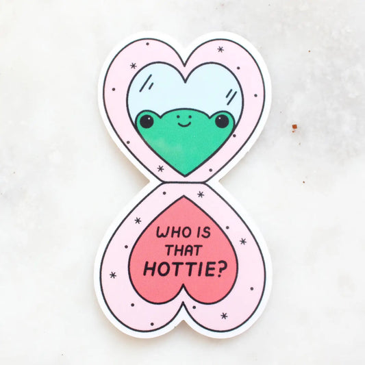 Die-cut sticker in the shape of a heart-shaped open makeup compact. Illustration of a frog looking into a mirror. "who is that hottie" in black text. 