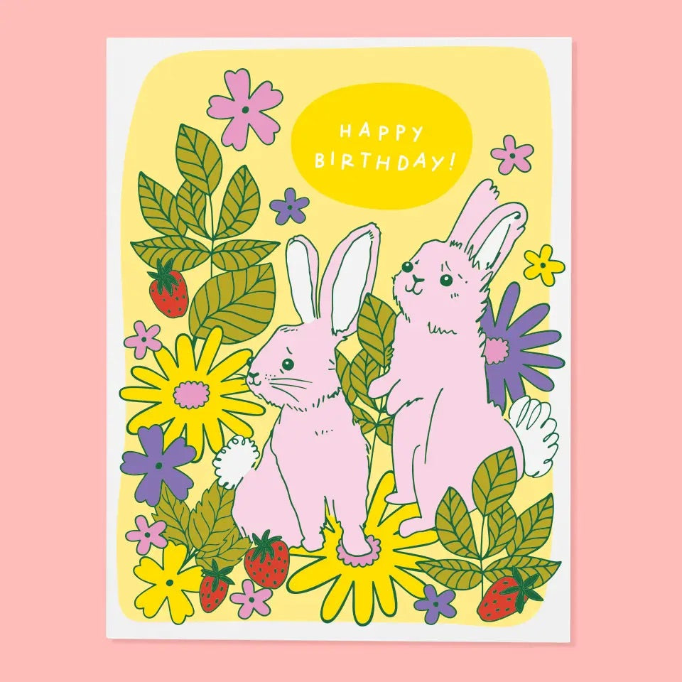 White greeting card with full color yellow woodland background. Illustration of two pink bunnies. White text “happy birthday” 