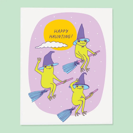 White card with purple background. Illustration of three green frogs on brooms wearing witches hats. Purple text reads “happy haunting!”