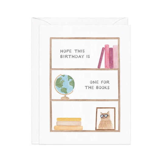 White card with an illustration of a bookshelf. Illustrations of books, a globe, and a framed picture of cat are on the shelves. Black text reads "hope this birthday is one for the books." 