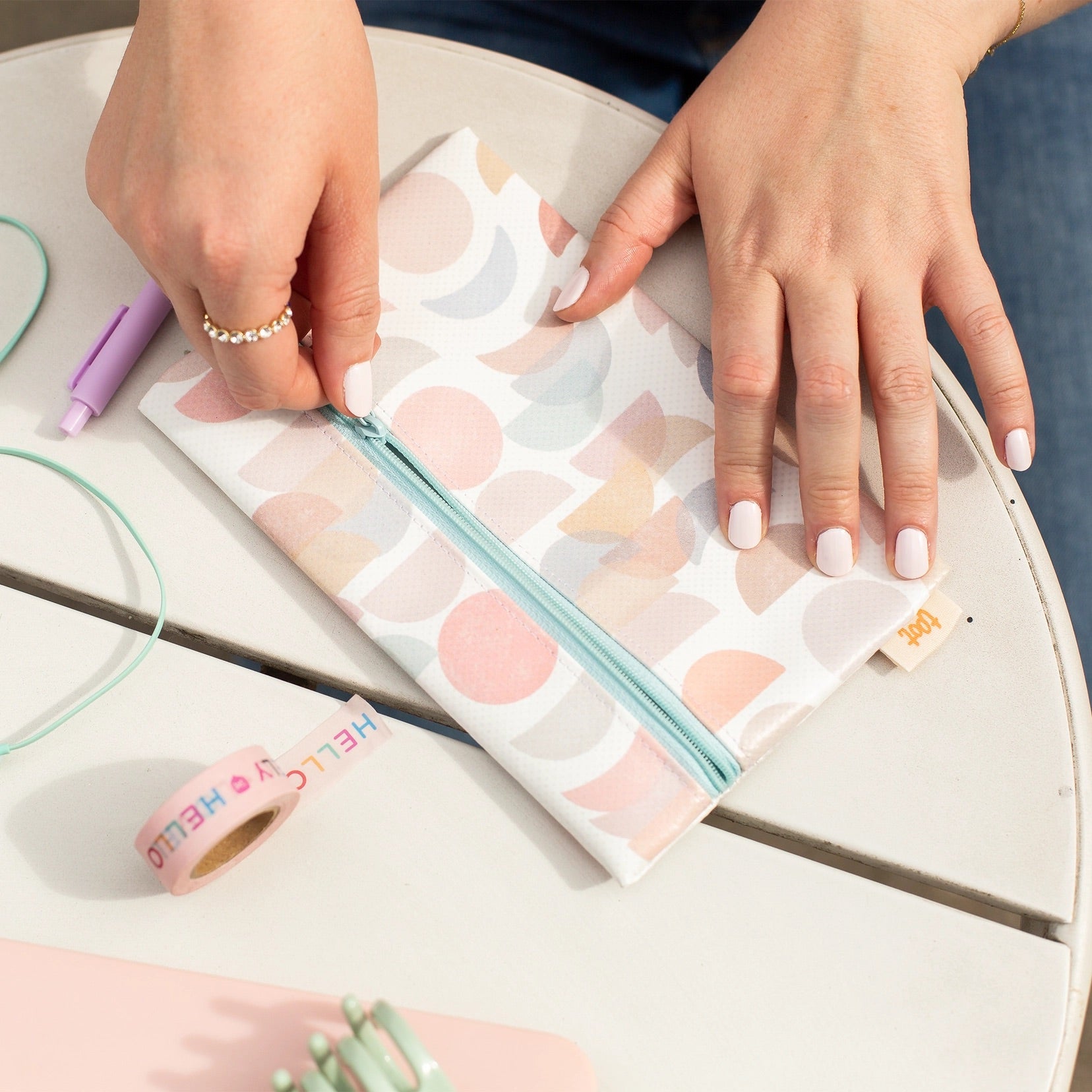 Two hands on top of a white zip pouch. One hand is unzipping the blue zipping. Pouch is decorated with muted blue, tan, and pink moon pattens. 