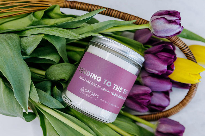 Clear jar filled with white candle. Purple label with text “tending to the tulips”