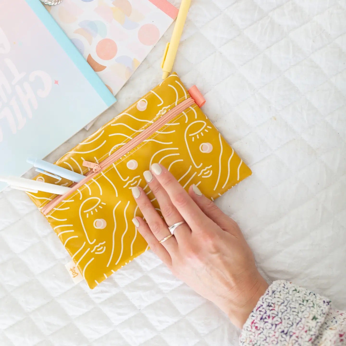 Hand touching a mustard colored pencil case. Abstract white squiggle design. 