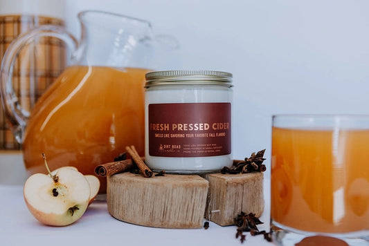 Clear jar filled with white candle. Brown label with gold text reading “fresh pressed cider.” Apple cider, cinnamon, and clove decor in background. 