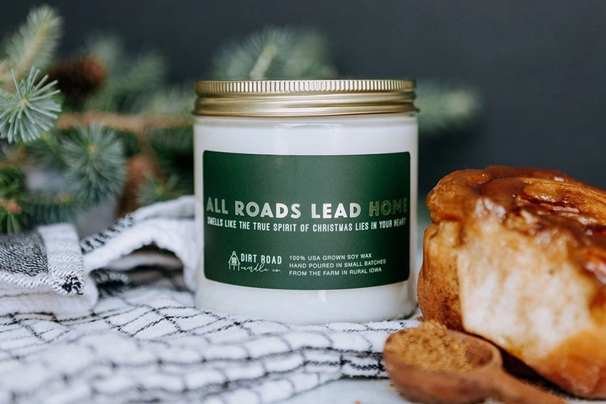 Clear jar filled with white candle. Green label with silver text “all roads lead home”