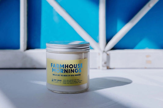 Clear jar filled with white candle. Yellow label with text “farmhouse mornings”