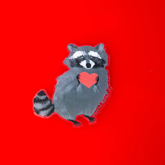 Die-cut sticker in the shape of a raccoon holding a heart. 