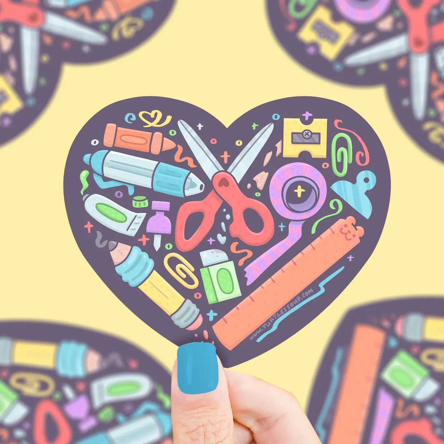 Hand holding a heart-shaped sticker with navy background and multicolor illustrations of school supplies. 