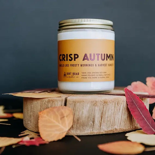 Clear jar with white candle. Orange label with dark red text reading "crisp autumn." Leaf and tree stump decor. 