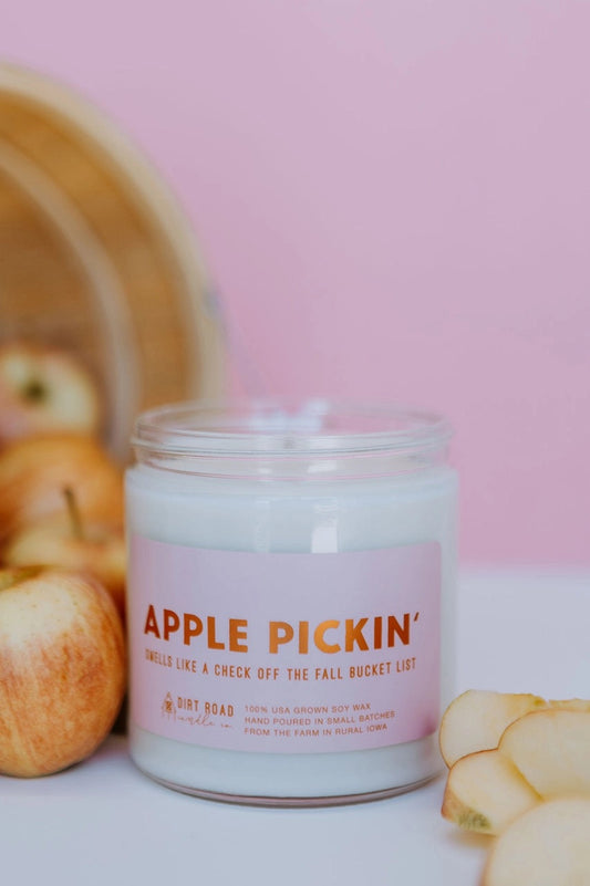 Clear jar with white candle. Pink label with red text reads "apple pickin'" Pink background with apple decor. 