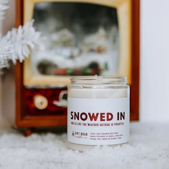 Clear jar filled with white candle. White label with red text “snowed in”