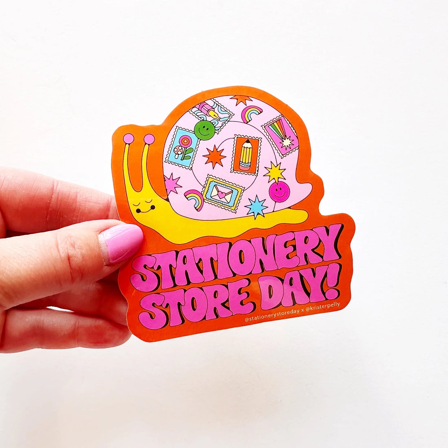 Hand holding a die-cut sticker. Orange background. Multicolor snail illustration with stamps on its shell. Pink bubble letter text reads "Stationery Store Day!" 