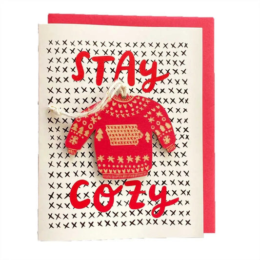 White greeting card with red text “stay cozy.” Sweater shaped red ornament is attached 