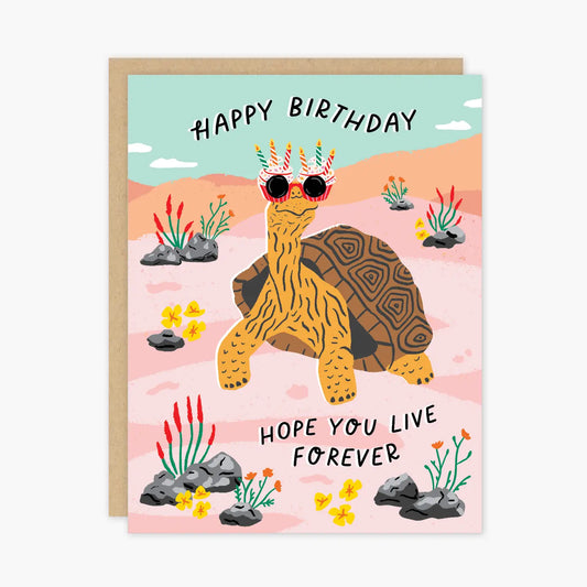 White card with an illustrated blue, tan, and red desert background. Illustration of a tortoise wearing red sunglasses with birthday candles on them. "Happy birthday. Hope you live forever" in black text. 