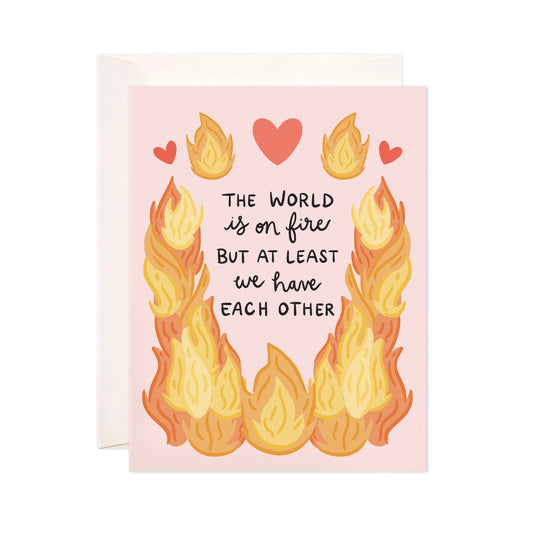 Card with flames and hearts design on a pink background. Black text: the world is on fire but at least we have each other 