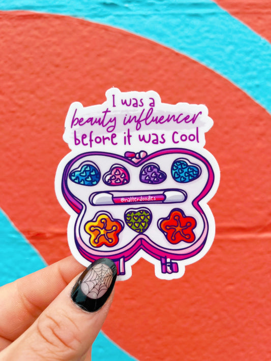 Hand holding a die-cut sticker in the shape of a 1990s kid's makeup palate. Pink text reads "I was a beauty influencer before it was cool" 