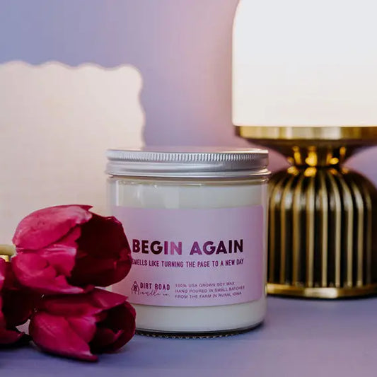 Candle jar with 'Begin Again' text next to pink roses on a soft purple background
