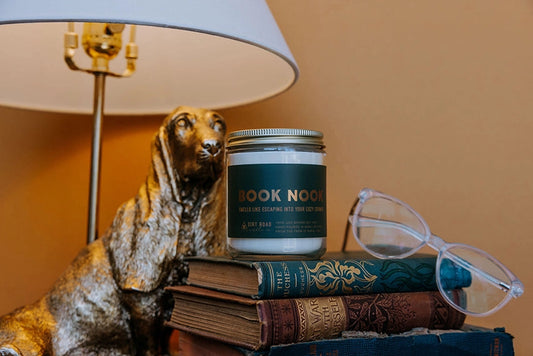 Candle labeled 'Book Nook' sitting on books with a lamp and glasses in the background