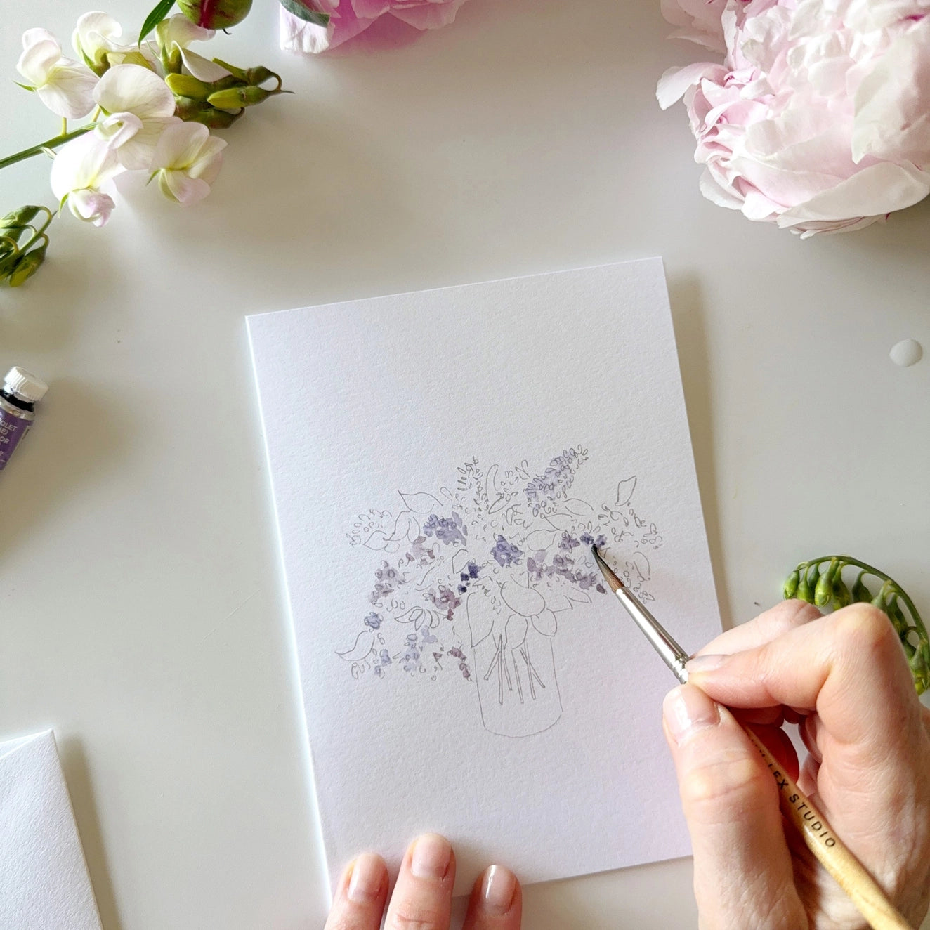 Person painting a floral design on a white card with flowers and art supplies around.