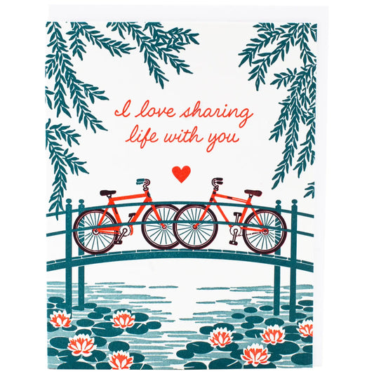 White greeting card with an illustration of two bikes on a bridge. Red text: I love sharing life with you 