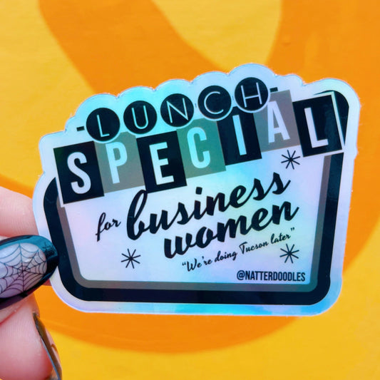 Hand holding a die-cut sticker with a holographic background. Sticker text reads "lunch special for business women, we're doing Tucson later." 