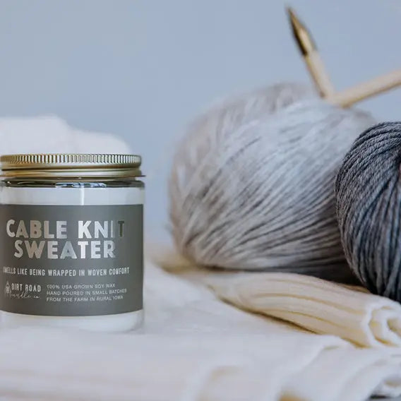 Candle labeled 'Cable Knit Sweater' with yarn and knitting needles in the background