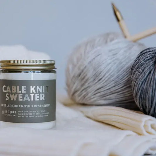 Candle labeled 'Cable Knit Sweater' with yarn and knitting needles in the background