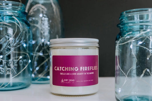 White candle in a clear jar. Purple label with text: catching fireflies 