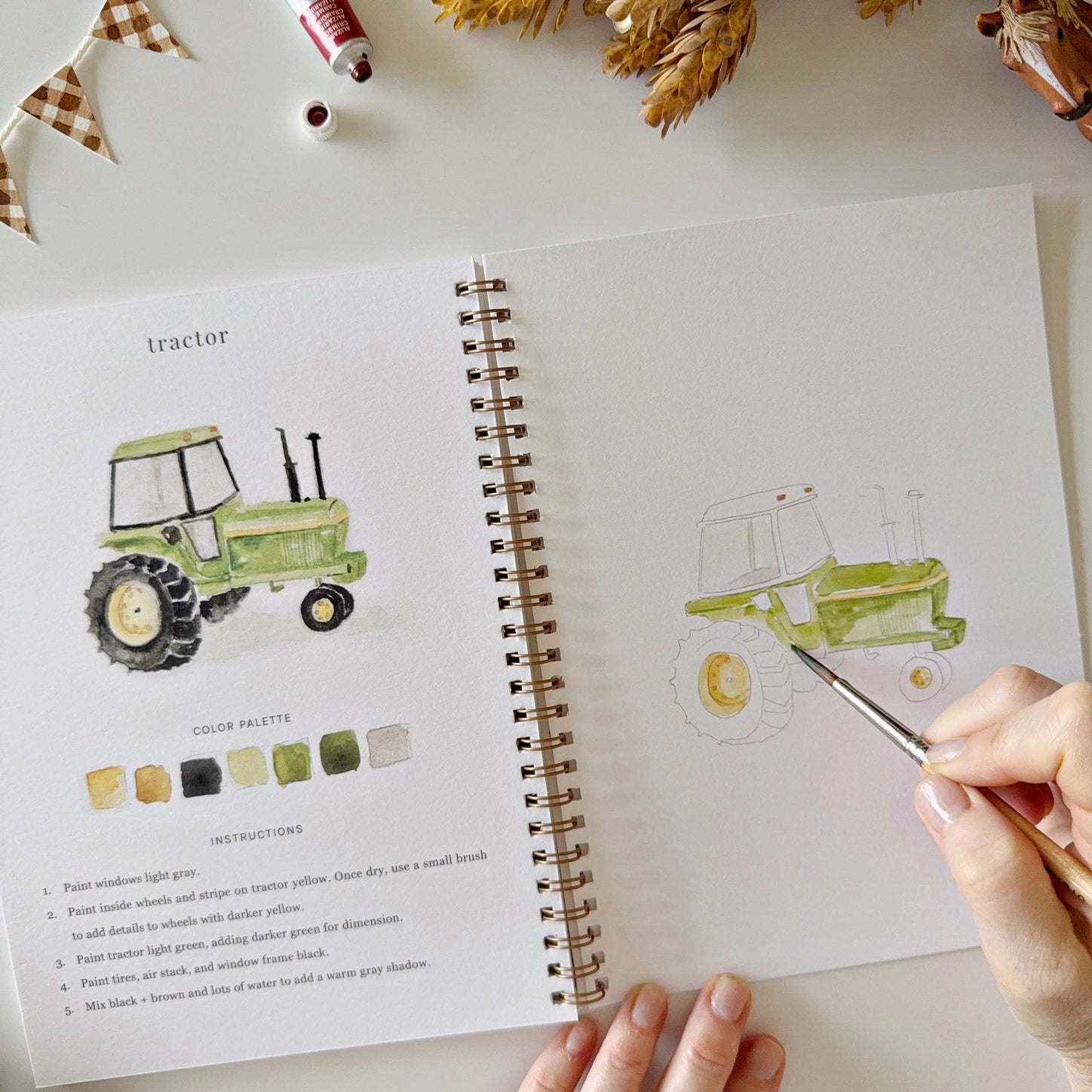 Person drawing a tractor from a watercolor book on a table with paint supplies.