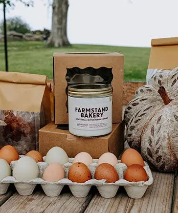 Jar candle labeled Farmstand Bakery next to eggs and a pumpkin 