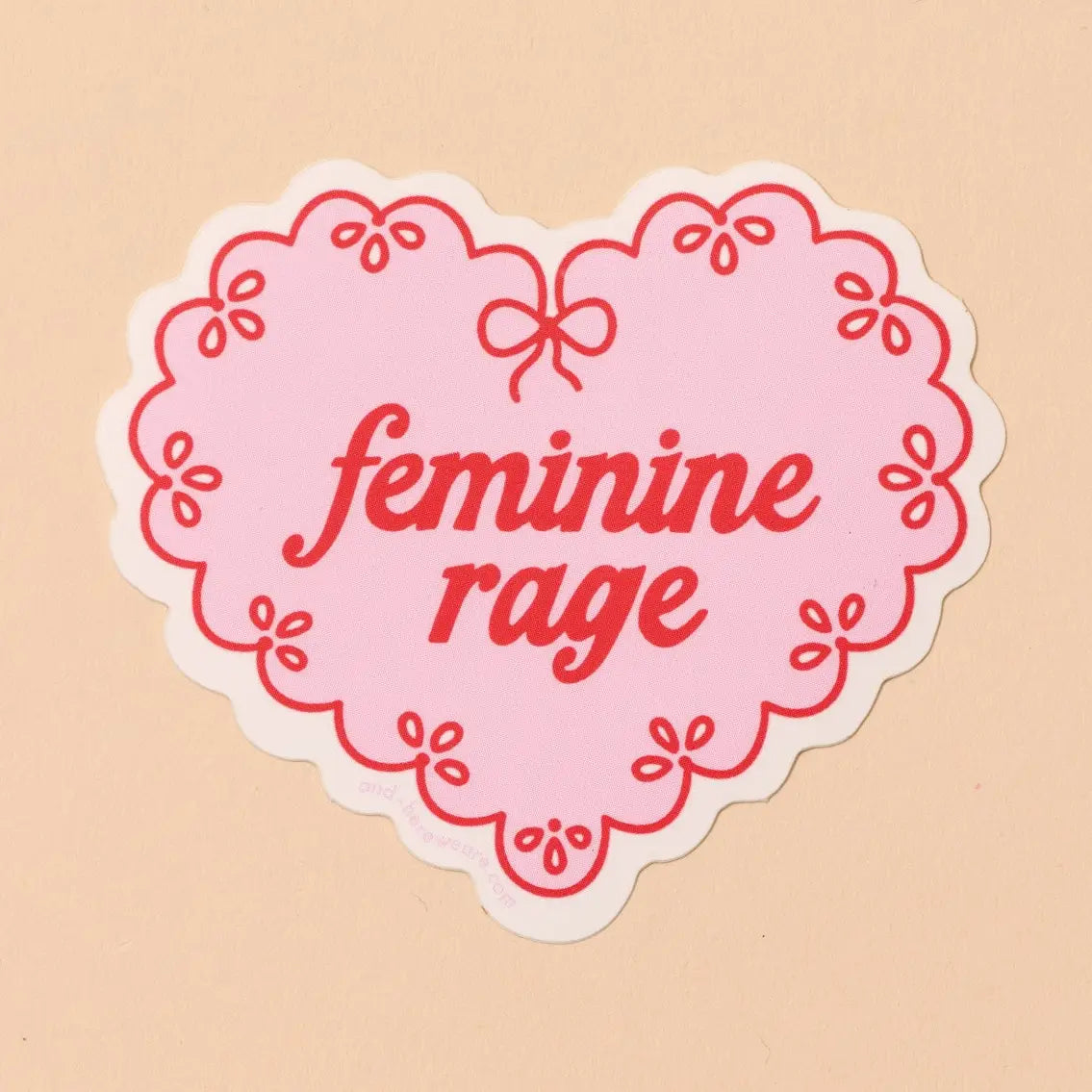 Pink and red heart-shaped sticker with 'feminine rage' text on a beige background