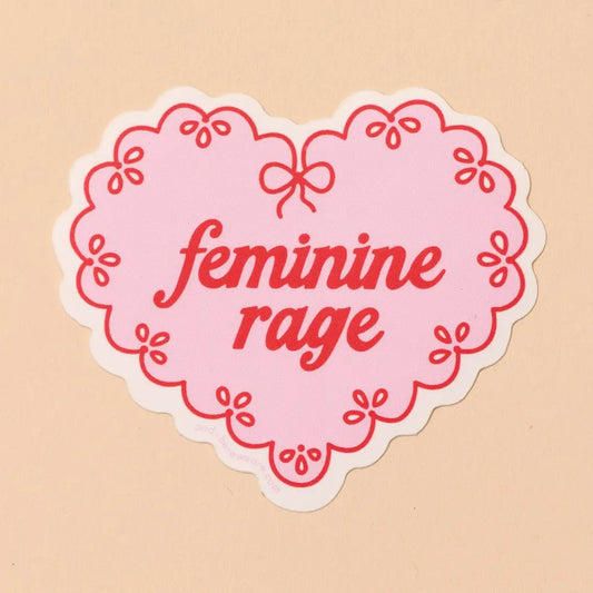 Pink and red heart-shaped sticker with 'feminine rage' text on a beige background