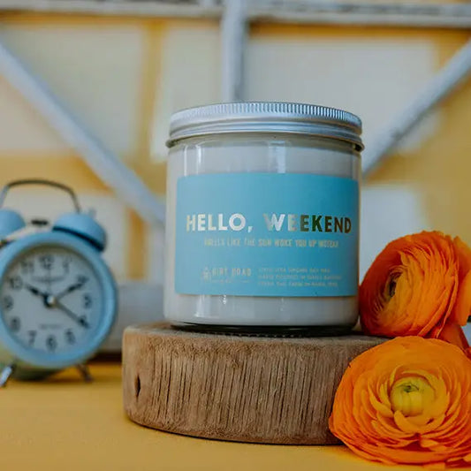 Candle labeled 'Hello, Weekend' with an alarm clock and flowers in the background