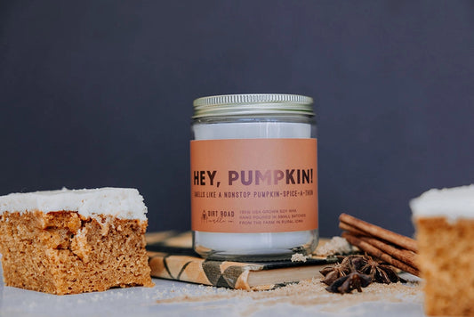 Clear jar filled with white candle. Orange label with dark orange text reads "hey, pumpkin!." Cinnamon sticks and pumpkin bar decor in the foreground. 