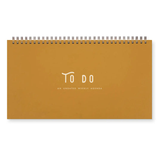 Front cover of top-spiral notebook. Mustard color with white text “to do, an undated weekly agenda” 