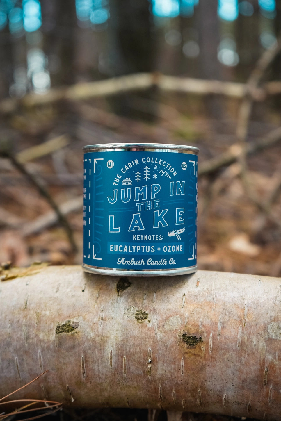 Blue candle canister labeled 'Jump in the Lake' on a log with a forest background