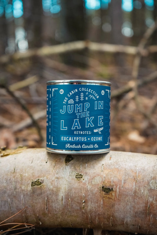 Blue candle canister labeled 'Jump in the Lake' on a log with a forest background