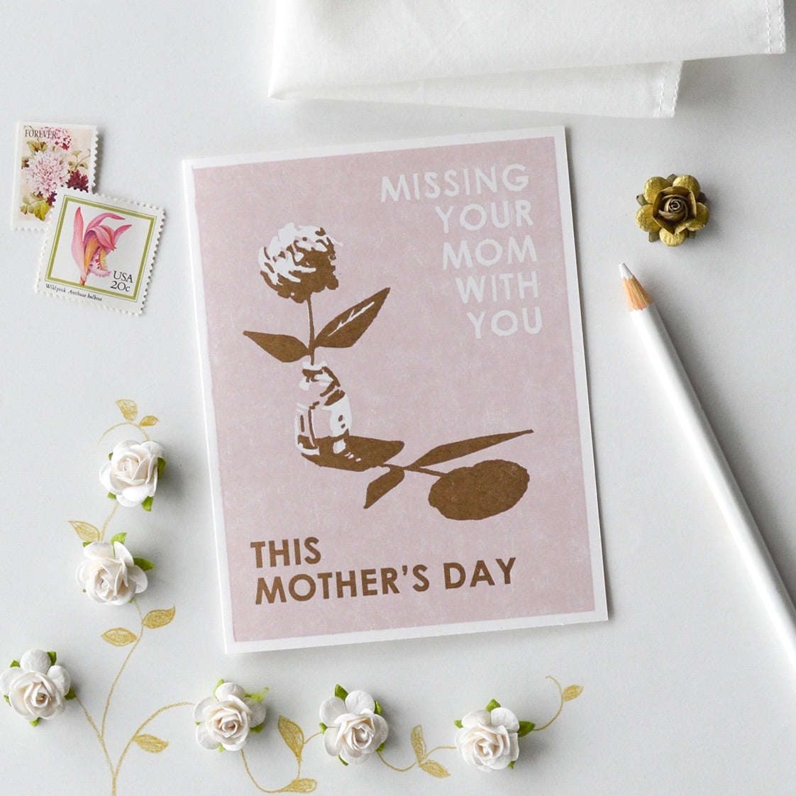 White card with full color pink background. White and brown floral print. White text reads "missing your mom with you" and brown text reads "this mother's day." Stamps, pencil, and floral decor around the card. 