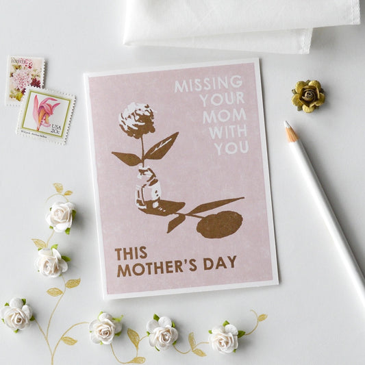 White card with full color pink background. White and brown floral print. White text reads "missing your mom with you" and brown text reads "this mother's day." Stamps, pencil, and floral decor around the card. 