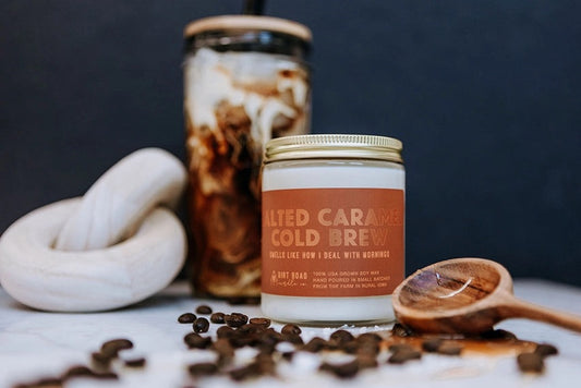 Clear jar filled with white candle. Light brown label with gold text reading "salted caramel cold brew." Coffee decor in the foreground and background