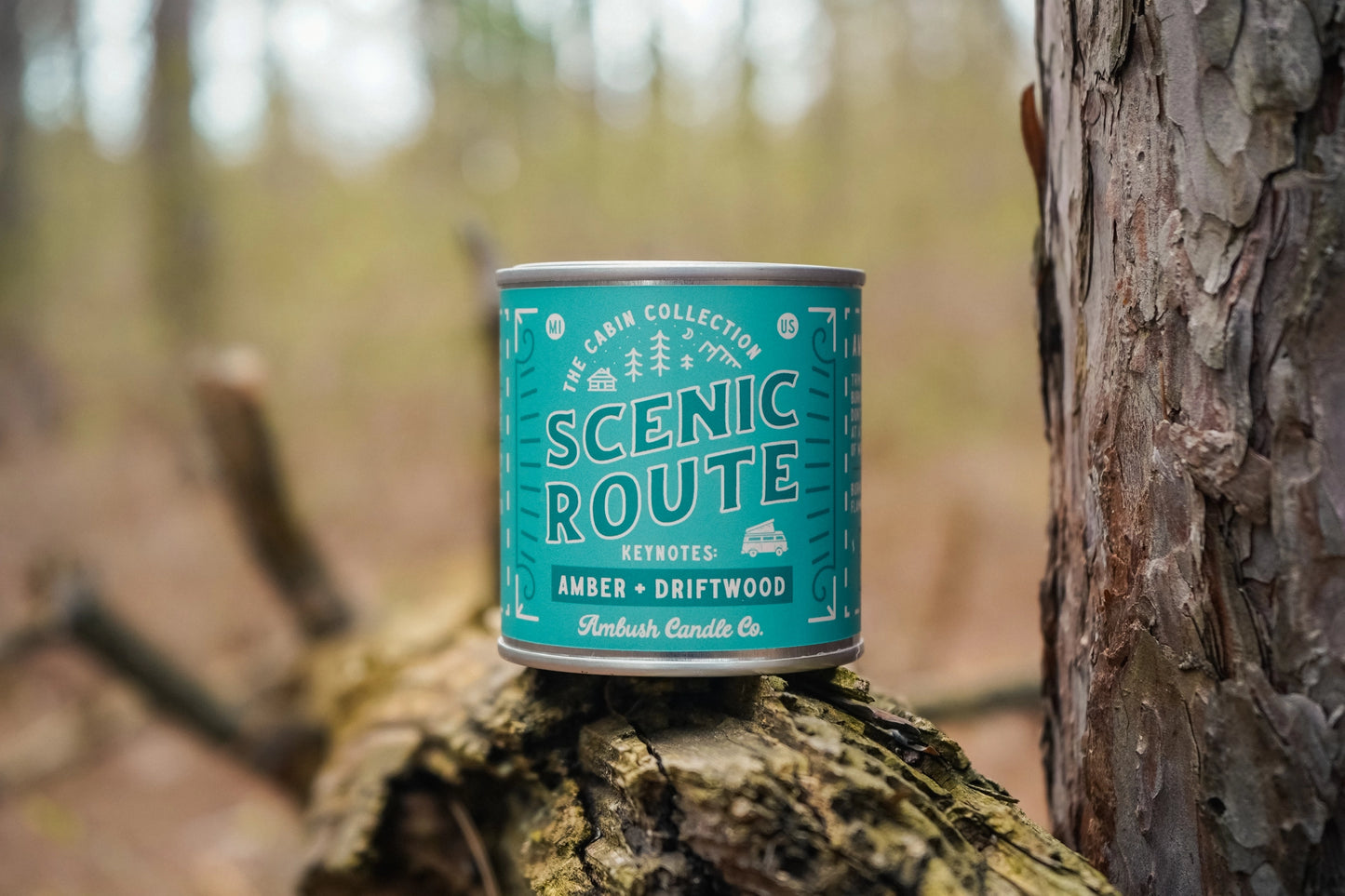Teal candle canister labeled 'Scenic Route' on a wooden log with a forest background