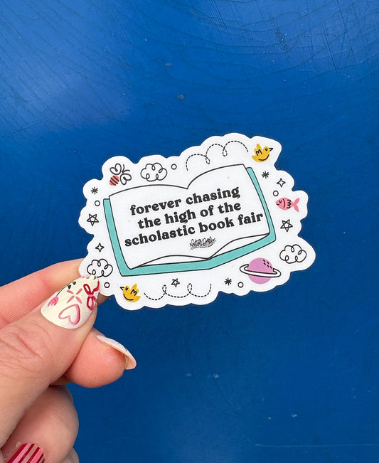 Sticker with text 'forever chasing the high of the scholastic book fair' held against a blue background